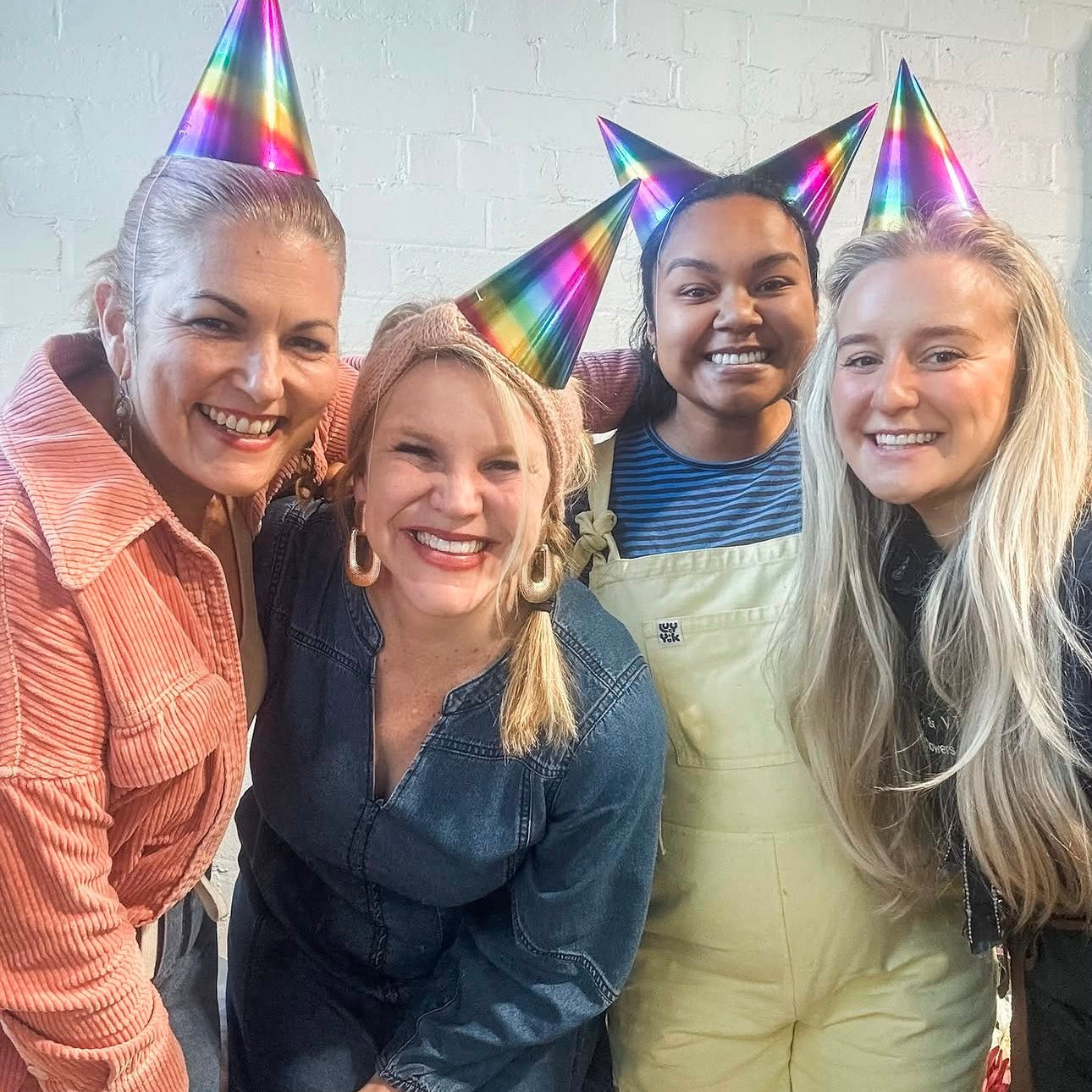 Four women smiling and posing together while wearing shiny rainbow party hats against a white brick wall.